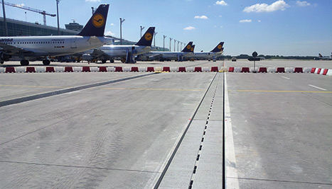 Eine Entwässerungsrinne führt den Blick entlang zu Flugzeugen in Parkpositionen, die vor einem Terminal stehen..